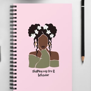 May include: A pink spiral bound notebook with a black and white illustration of a woman with flowers in her hair. The text "shaping my fro & behavior" is printed on the notebook.