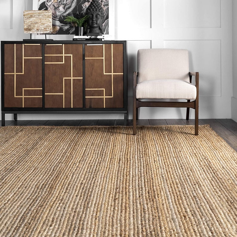 May include: A natural fiber rug with a woven texture in a light brown color. The rug is placed in a living room setting with a wooden cabinet and a chair.