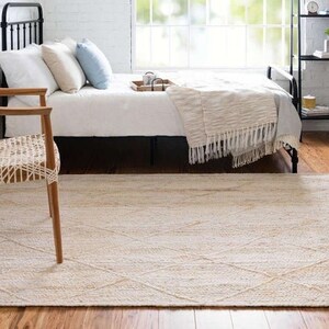 May include: A light beige woven rug with a diamond pattern, placed in a bedroom with a bed and a wooden chair.