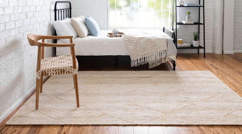May include: A light beige woven rug with a diamond pattern, placed in a bedroom with a bed and a wooden chair.