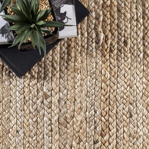 May include: A natural fiber braided rug with a light brown color. The rug is made of tightly woven strands of jute, creating a textured and durable surface.