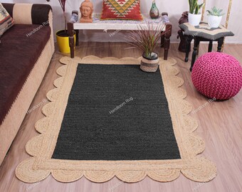 May include: A rectangular jute rug with a dark gray center and a natural tan scalloped border. The rug is placed on a light wood floor, with a pink knitted pouf and other decorative items in the background.