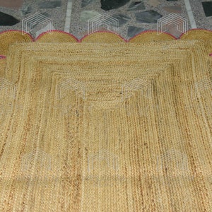May include: A natural fiber rug with a square shape and a scalloped border. The rug is made of jute and has a light brown color. The border is a light pink color.