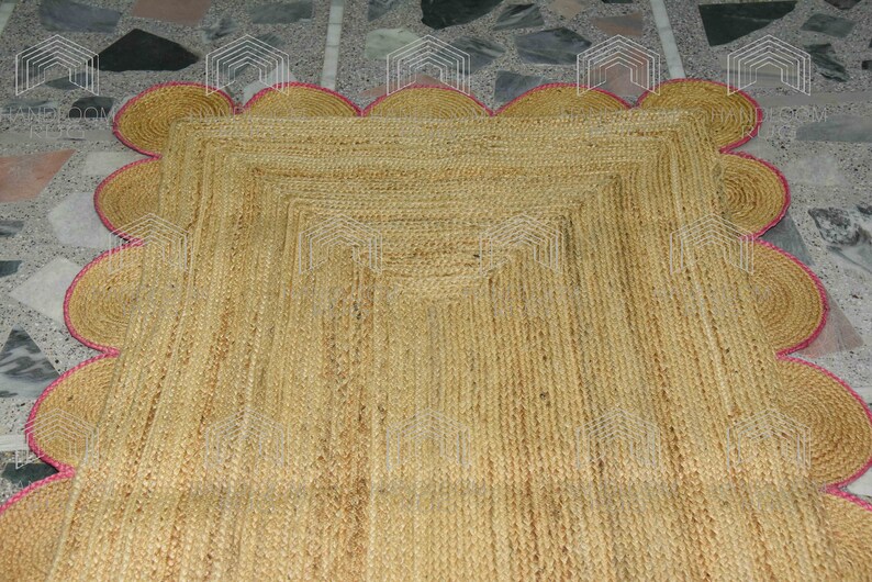 May include: A natural fiber rug with a square shape and a scalloped border. The rug is made of jute and has a light brown color. The border is a light pink color.