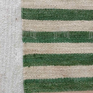 Op de afbeelding: Close-up van een groen-wit gestreept tapijt. Het tapijt is gemaakt van een geweven materiaal en heeft een natuurlijke, rustieke uitstraling.