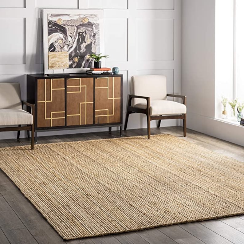 May include: A natural fiber rug in a light brown color with a woven texture. The rug is placed in a living room setting with two chairs and a cabinet.