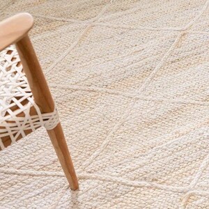 May include: A close-up of a white woven rug with a diamond pattern. The rug is made of natural fibers and has a slightly textured surface. A wooden chair leg is visible in the bottom right corner of the image.