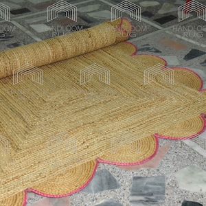 May include: A natural fiber rug with a square shape and a scalloped edge. The rug is made of woven jute and has a pink trim.
