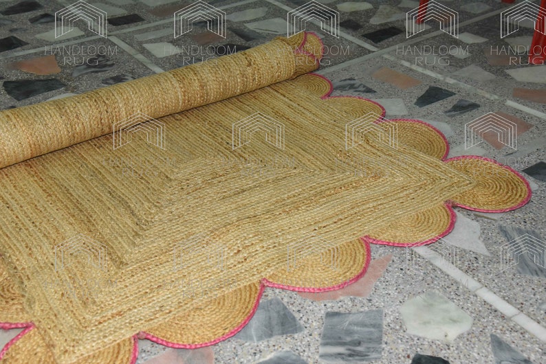 May include: A natural fiber rug with a square shape and a scalloped edge. The rug is made of woven jute and has a pink trim.