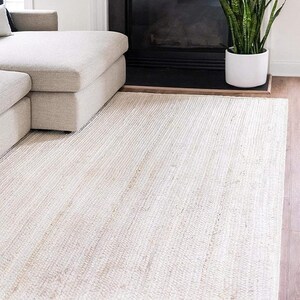 May include: A white jute rug with a subtle woven texture. The rug is laid out on a hardwood floor in a living room setting.
