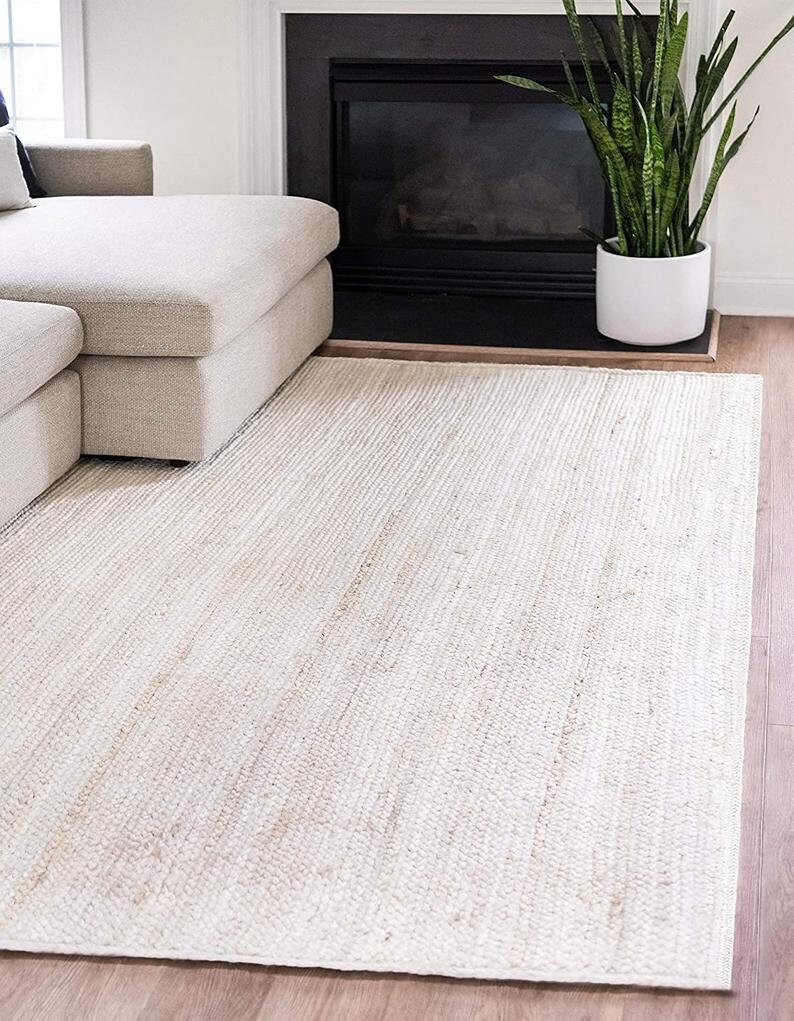 May include: A white jute rug with a subtle woven texture. The rug is laid out on a hardwood floor in a living room setting.