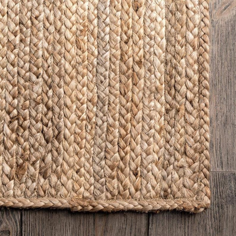 May include: Close-up of a natural fiber rug with a woven texture. The rug is made of braided jute and has a light brown color.