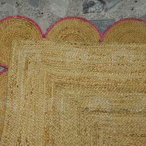 May include: A natural fiber rug with a square shape and a scalloped border. The rug is made of jute and has a light brown color. The border is a light pink color.