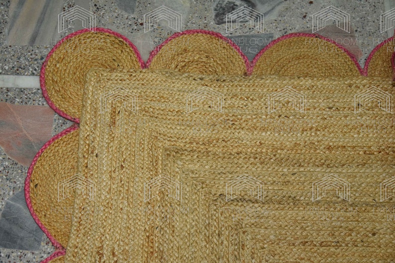 May include: A natural fiber rug with a square shape and a scalloped border. The rug is made of jute and has a light brown color. The border is a light pink color.