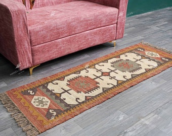 May include: A long, rectangular rug with a geometric pattern in shades of red, brown, beige, and white. The rug has fringe on both ends and is made of natural fibers.
