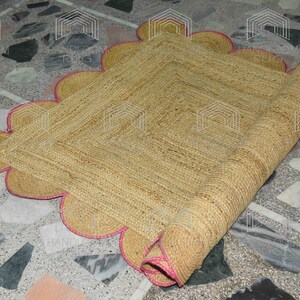 May include: A natural fiber rug with a scalloped edge and pink trim. The rug is rolled up and partially unrolled, revealing the woven texture.