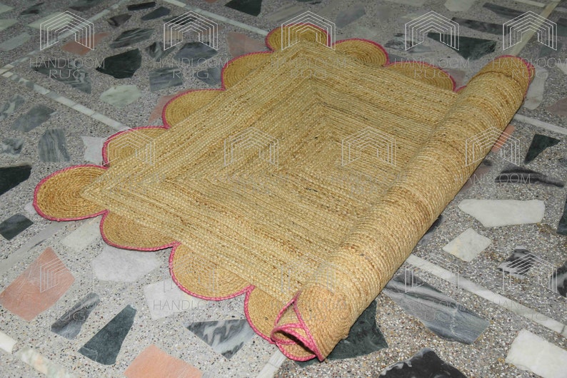 May include: A natural fiber rug with a scalloped edge and pink trim. The rug is rolled up and partially unrolled, revealing the woven texture.