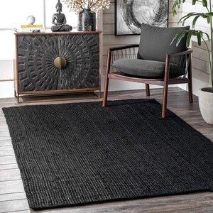 May include: Black jute rug with a woven texture. The rug is placed in a living room setting with a wooden chair and a cabinet with a decorative design.