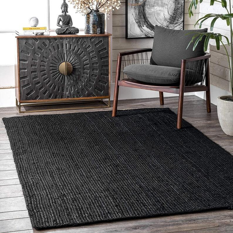May include: Black jute rug with a woven texture. The rug is placed in a living room setting with a wooden chair and a cabinet with a decorative design.