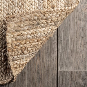 May include: Close-up of a natural fiber rug with a woven texture. The rug is a light brown color and is folded over on itself, revealing the underside of the weave. The rug is lying on a wood floor.