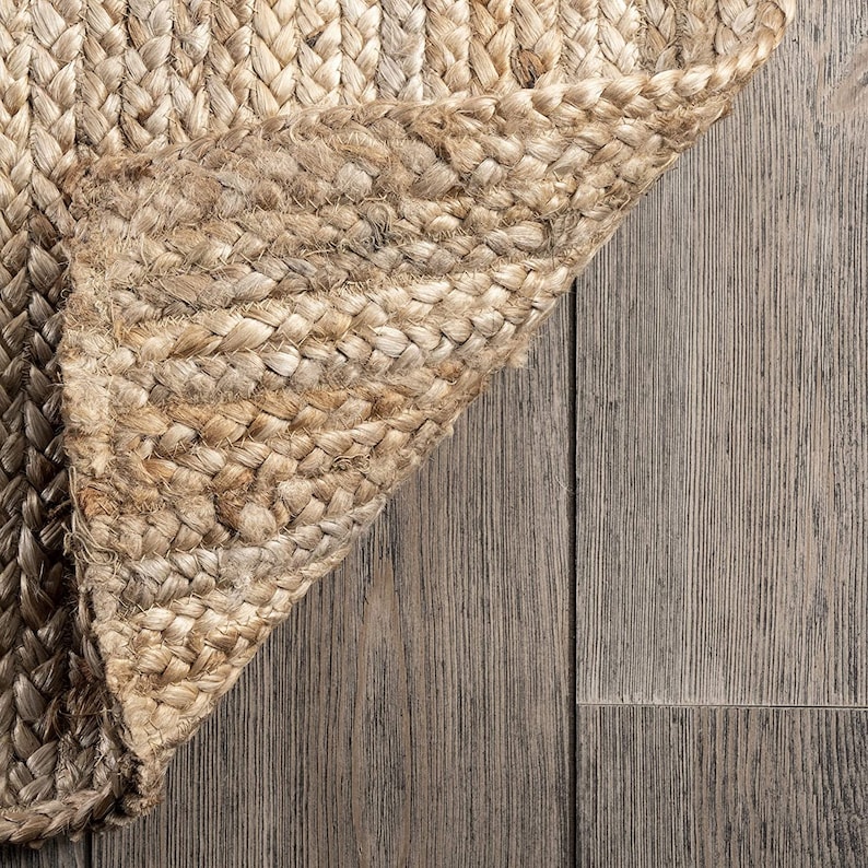May include: Close-up of a natural fiber rug with a woven texture. The rug is a light brown color and is folded over on itself, revealing the underside of the weave. The rug is lying on a wood floor.
