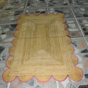 May include: A natural fiber rug with a pink scalloped border. The rug is made of jute and has a natural, earthy color. The rug is laid out on a tiled floor.