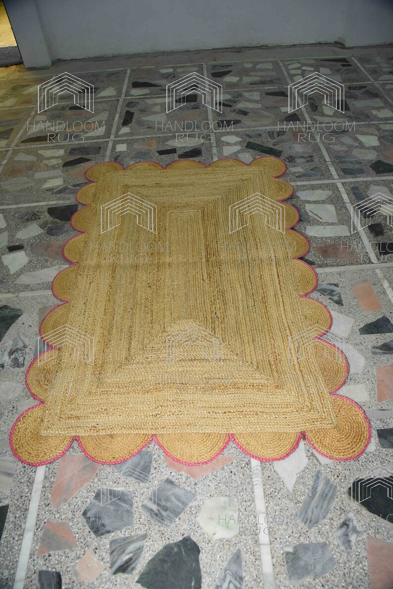 May include: A natural fiber rug with a pink scalloped border. The rug is made of jute and has a natural, earthy color. The rug is laid out on a tiled floor.