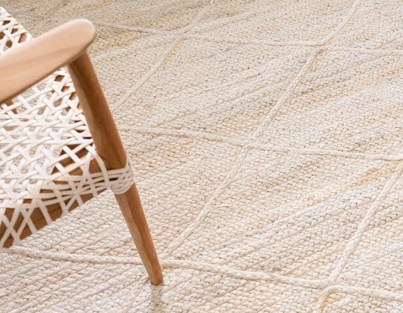May include: A close-up of a white woven rug with a diamond pattern. The rug is made of natural fibers and has a soft, textured surface. A wooden chair leg is visible in the bottom right corner of the image.