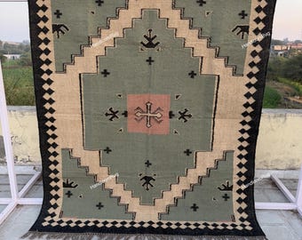 May include: A rectangular rug with a geometric design. The rug features a central sage green field with a pink square and black cross-like shapes. The border is tan with black diamond shapes and a fringe along the bottom. The rug is displayed outdoors.