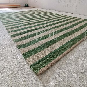 Op de afbeelding: Een groen en natuurlijk jute gestreept tapijt. Het tapijt is handgeweven en heeft een natuurlijke, rustieke uitstraling. Het tapijt is perfect om elke kamer een vleugje natuurlijke schoonheid te geven.