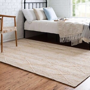 May include: A cream-colored woven rug with a diamond pattern. The rug is placed in a bedroom with a wooden chair and a bed with a white blanket and pillows.