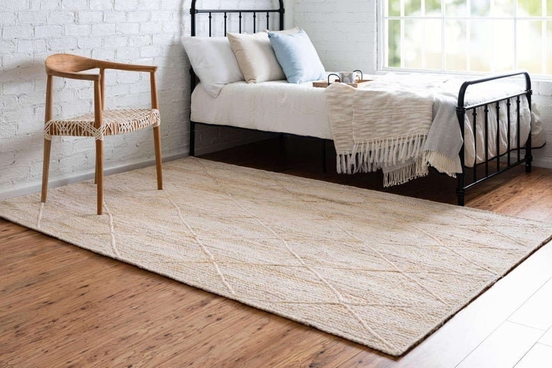 May include: A cream-colored woven rug with a diamond pattern. The rug is placed in a bedroom with a wooden chair and a bed with a white blanket and pillows.