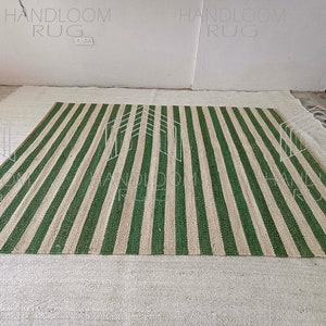 Op de afbeelding: Een handgeweven tapijt met groene en beige strepen. Het tapijt is gemaakt van natuurlijke vezels en heeft een rustieke uitstraling.