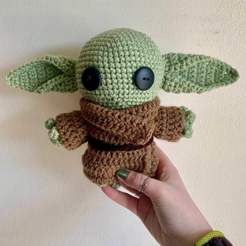 May include: A crocheted green and brown plush toy resembling a character from the Star Wars franchise. The toy has large eyes and a brown crocheted robe.