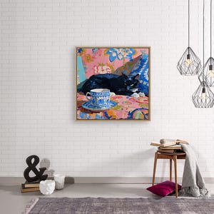 May include: A framed painting depicts a black cat resting on a floral patterned surface. A blue and white teacup and saucer sit in the foreground. The artwork is set against a white brick wall, with decorative hanging lights.