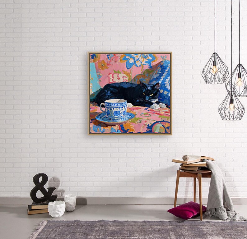 May include: A framed painting depicts a black cat resting on a floral patterned surface. A blue and white teacup and saucer sit in the foreground. The artwork is set against a white brick wall, with decorative hanging lights.