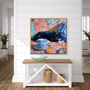 May include: A framed painting depicts a black cat resting on a floral patterned surface. A blue and white teacup and saucer are in the foreground. The artwork has a pink, blue, and yellow color palette, with a wooden frame.
