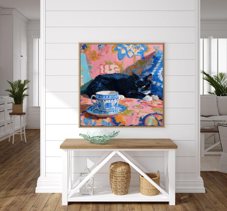 May include: A framed painting depicts a black cat resting on a floral patterned surface. A blue and white teacup and saucer are in the foreground. The artwork has a pink, blue, and yellow color palette, with a wooden frame.