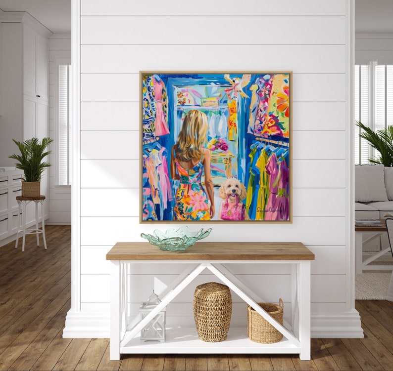 Puede incluir: Una pintura enmarcada representa a una mujer con un vestido floral colorido, frente a un armario lleno de ropa vibrante. Un perro peque&ntilde;o con un atuendo rosa est&aacute; a su lado. La obra de arte se exhibe sobre una consola de madera.