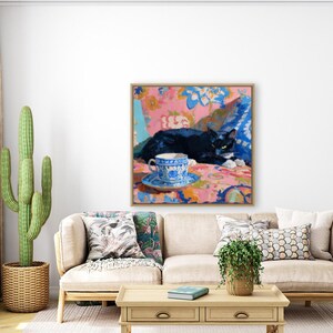 May include: A framed painting depicts a black cat resting on a patterned surface, with a blue and white teacup and saucer in the foreground. The artwork features a vibrant color palette of pink, blue, and white, creating a cozy and artistic scene.