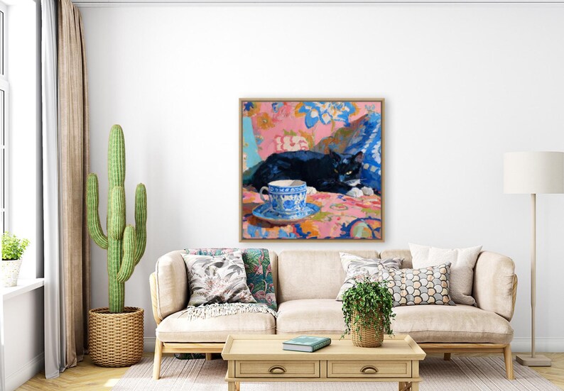 May include: A framed painting depicts a black cat resting on a patterned surface, with a blue and white teacup and saucer in the foreground. The artwork features a vibrant color palette of pink, blue, and white, creating a cozy and artistic scene.