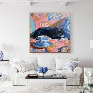 May include: An oil painting depicts a black cat resting on a patterned surface, with a blue and white teacup and saucer in the foreground. The artwork is framed and hangs above a white sofa, creating a cozy, artistic interior.