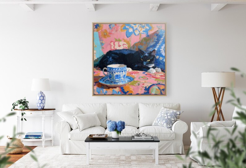 May include: An oil painting depicts a black cat resting on a patterned surface, with a blue and white teacup and saucer in the foreground. The artwork is framed and hangs above a white sofa, creating a cozy, artistic interior.