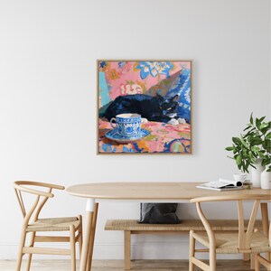 May include: A framed painting depicts a black cat resting on a floral patterned surface. A blue and white teacup and saucer sit in the foreground. The artwork features a vibrant color palette with pink, blue, and orange hues.