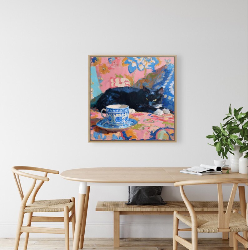 May include: A framed painting depicts a black cat resting on a floral patterned surface. A blue and white teacup and saucer sit in the foreground. The artwork features a vibrant color palette with pink, blue, and orange hues.