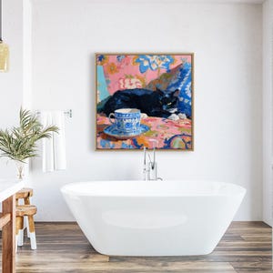 May include: A framed painting depicts a black cat resting on a colorful floral patterned surface. A blue and white teacup and saucer are in the foreground. The artwork is displayed in a modern bathroom setting with a white bathtub.