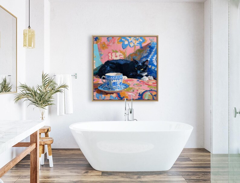 May include: A framed painting depicts a black cat resting on a colorful floral patterned surface. A blue and white teacup and saucer are in the foreground. The artwork is displayed in a modern bathroom setting with a white bathtub.