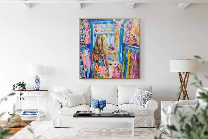 Puede incluir: Una pintura colorida representa a una mujer con un vestido floral mirando dentro de un armario lleno de ropa. Un perro con una camisa rosa est&aacute; a su lado. La obra de arte se exhibe sobre un sof&aacute; blanco en una sala de estar luminosa.