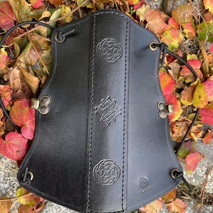 Könnte beinhalten: Schwarzer Leder-Bogenschießarmguard mit drei keltischen Knotendesigns und einem Riemenverschluss. Der Armguard liegt auf einem Bett aus Herbstlaub.