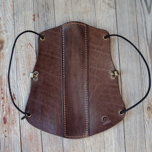 Peut inclure: Un protège-bras en cuir marron avec une sangle et une fermeture à boucle. Le protège-bras est doté d'un design cousu et d'un petit logo en bas.